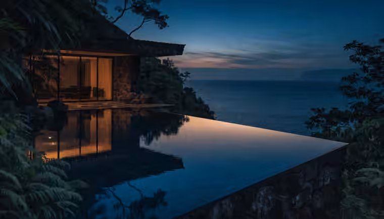 A cliffside villa lit from within above the ocean at dusk